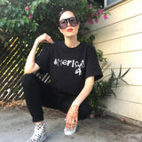 Leah Deluxe wearing a black tee-shirt that has America spelled with a bullet-pierced arrowhead, shackles, a noose, handgun, bomb, handcuff and hand, and a third-eye pyramid