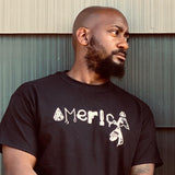 Man wearing a black tee-shirt that has America spelled with a bullet-pierced arrowhead, shackles, a noose, handgun, bomb, handcuff and hand, and a third-eye pyramid